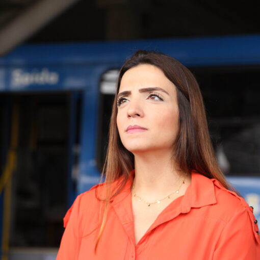 No primeiro plano, a vereadora Marcela Trópia em estação de ônibus olhando para o horizonte. No segundo plano, há um ônibus do transporte coletivo de BH desfocado. A vereadora veste camisa laranja.