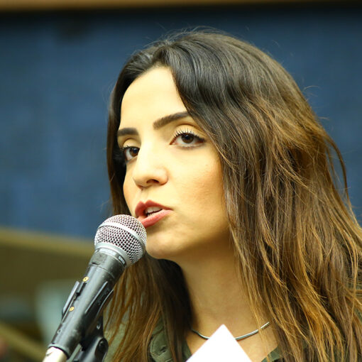 Marcela Trópia em primeiro plano falando com o microfone parecendo