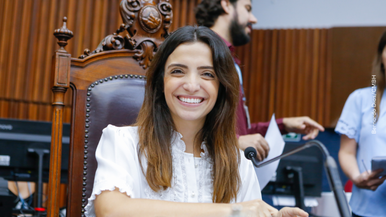Marcela Trópia sorrindo para a câmera sentada na cadeira da presidência com blusa de renda branca