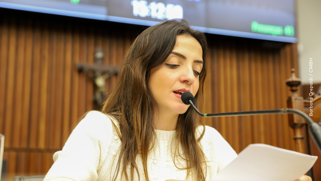 Vereadora Marcela Trópia sentada falando ao microfone. Atrás tem uma cruz e um pedaço do telão do Plenário
